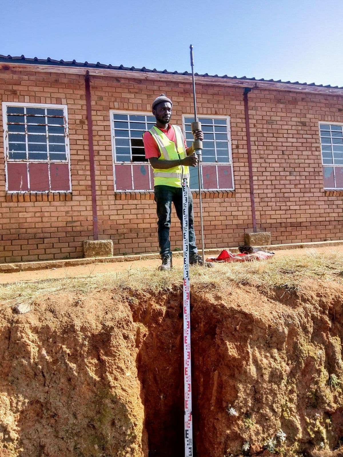 Worker holding a measuring staff at the edge of a deep excavation beside a brick building.