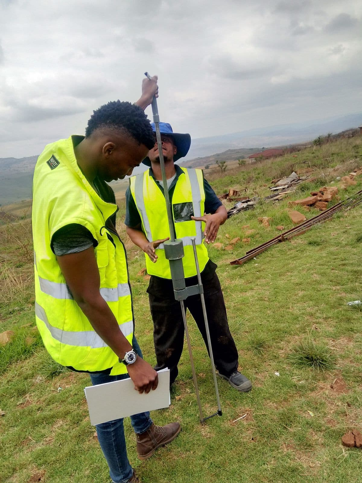 Two team members in reflective vests carrying out geotechnical field testing on open ground.
